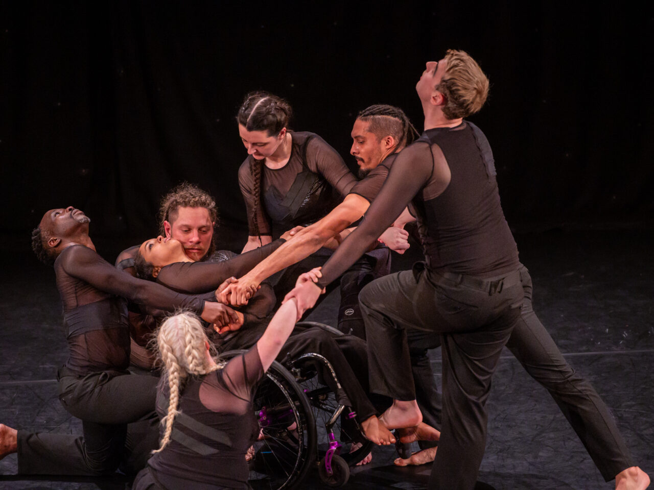 All seven dancers are connected holding hands. Arms are overlapped, some are stood up, some on the floor and in-between, all looking different ways. The wheelchair user is in the centre falling backwards into into another dancer.