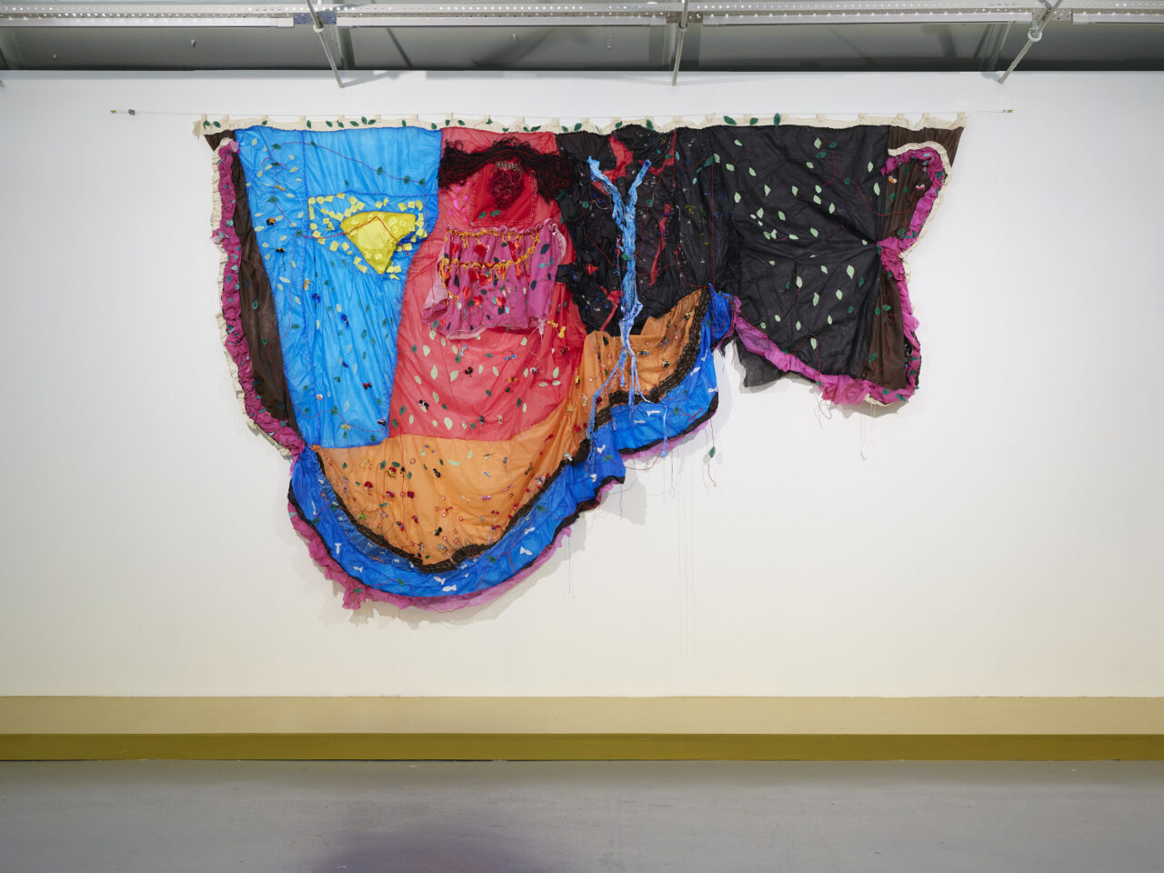 Hanging colourful textile work. The works are made up of materials including mixed fabrics, coal ink, imitation jewellery, stones, synthetic pearls, ribbons, buttons, straw, glitter and glue.