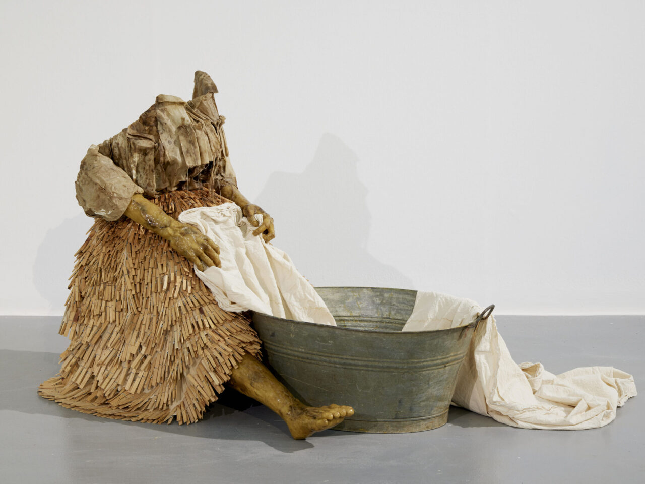 A mixed media sculpture of a woman seated holding onto a a piece of fabric, that is coming out of a tin bath. The woman's clothing has a skirt made out of wooden pegs, with a loose fitted top.
